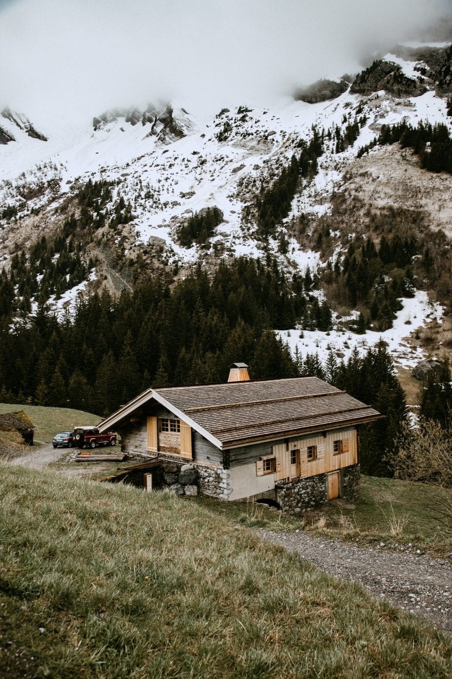 a rustic wooden house on a mountainside overlooking snowy mountains on a cloudy day