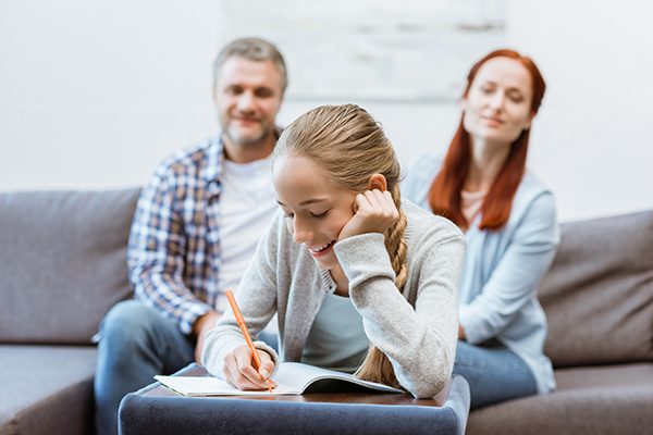 Teenage girl doing homework while parents resting on sofa at home