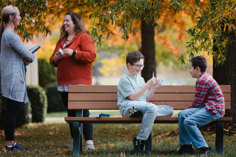 two homeschool moms chat outside while two boys play on a park bench