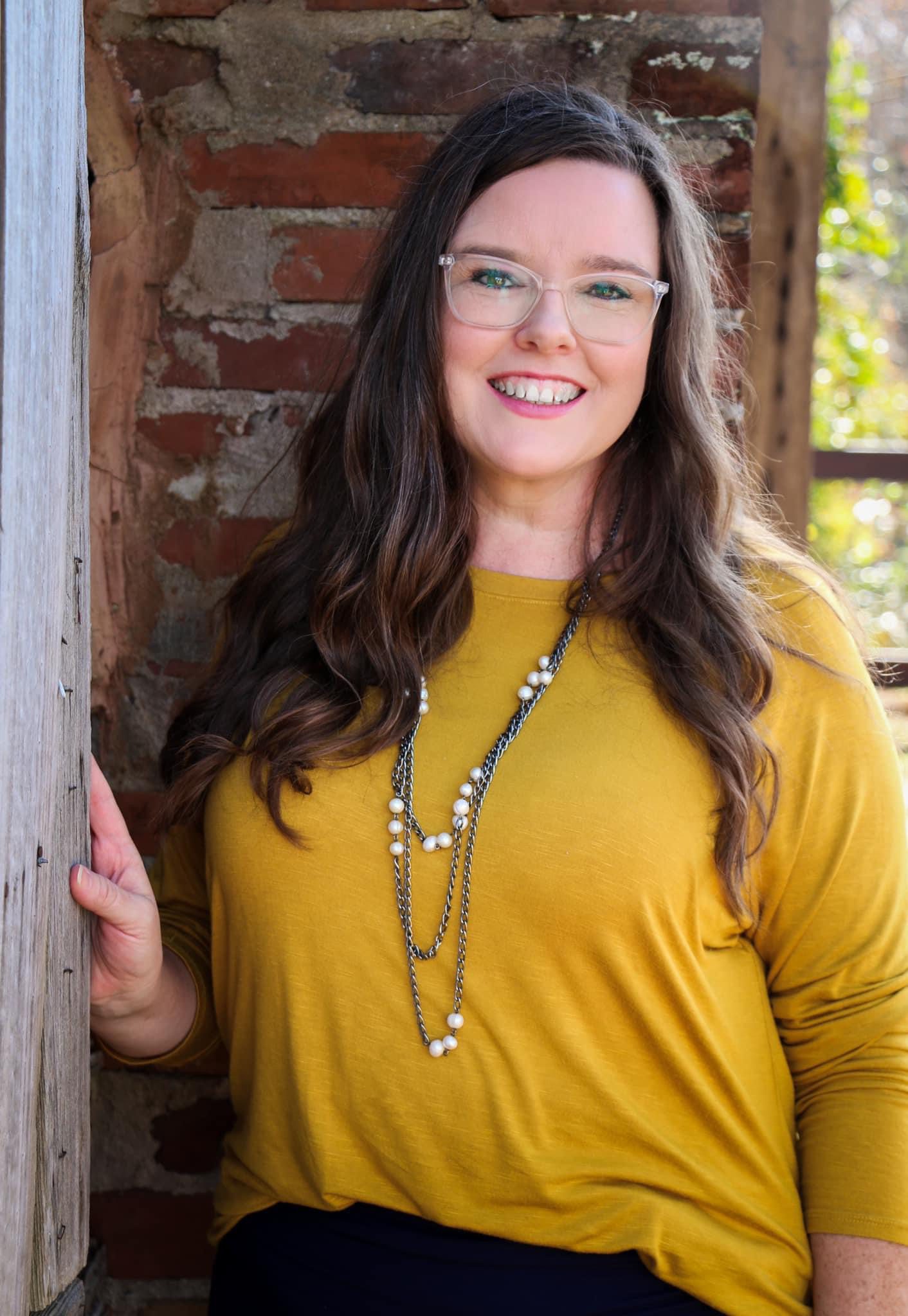 Woman in yellow shirt standing against a brick wall who works for Classical Conversations