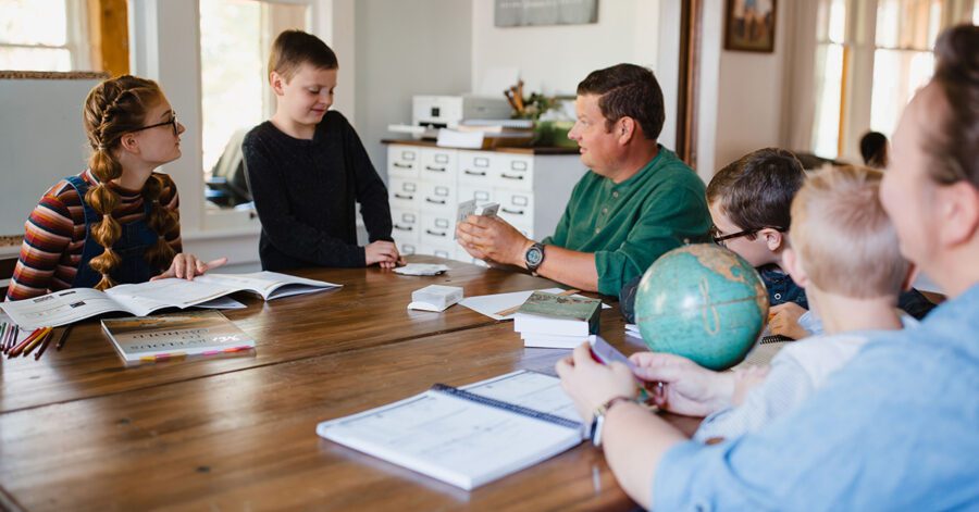 A family homeschool setting with children of different ages gathered around a table, engaging in interactive learning with books and educational tools, led by an adult.