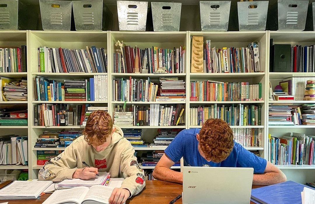 Two homeschool highschool boys working at a table with a bookshelf full of books behind them.