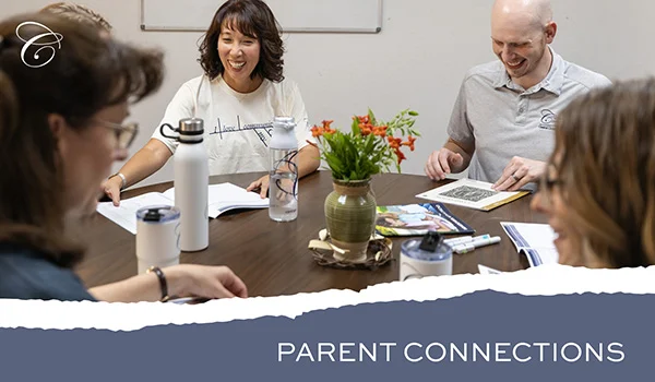 Classical Conversations parents and Director meeting together around a table, smiling and discussing educational materials with notebooks and flowers on the table
