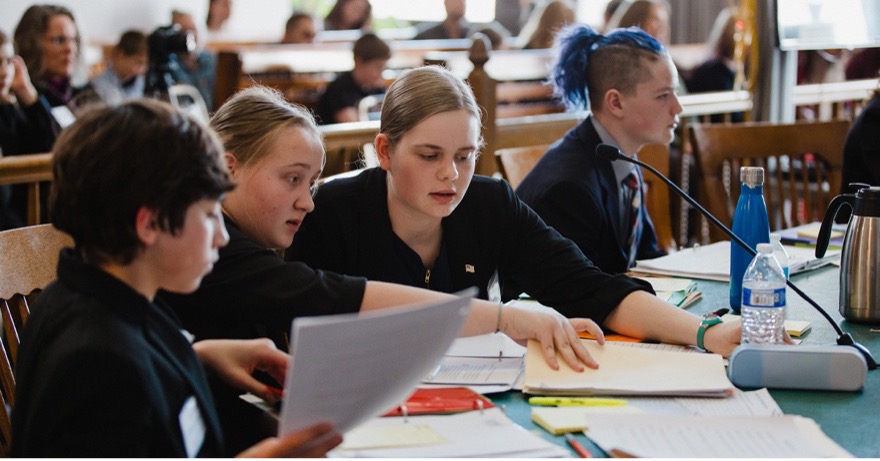 Challenge B students in formal attire conferring over case notes at a courtroom counsel table during Mock Trial
